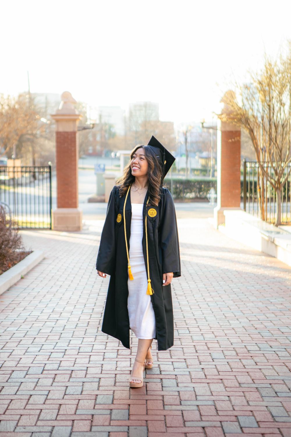 A smiling graduate in black cap and gown with gold tassel walks on a brick path outside a university campus.