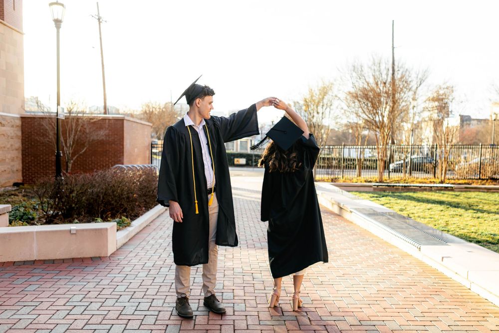 Two graduates in black caps and gowns dance together on a brick pathway during an outdoor graduation photo session.