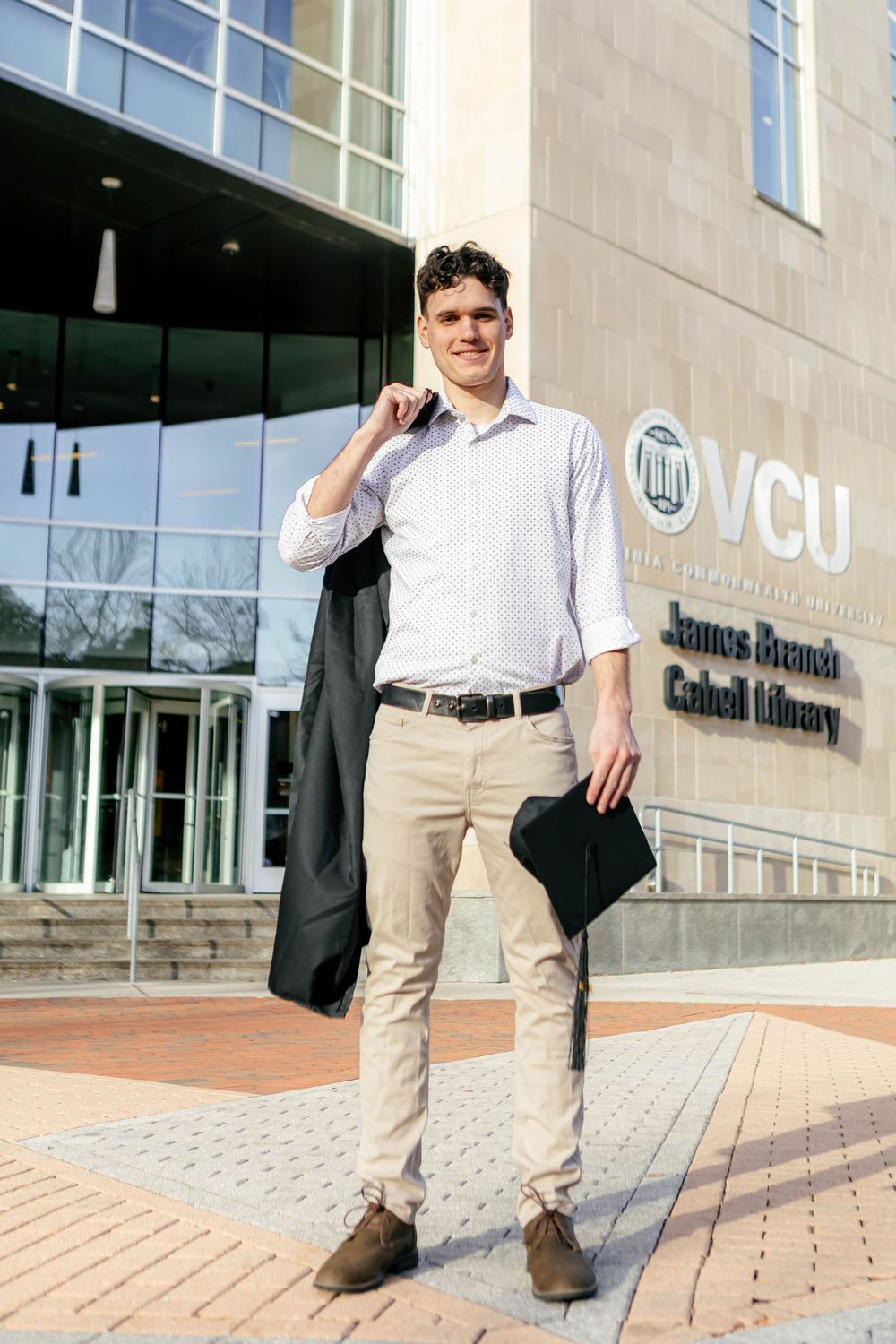 Young man in white shirt holding graduation gown and cap outside VCU James Branch Cabell Library.