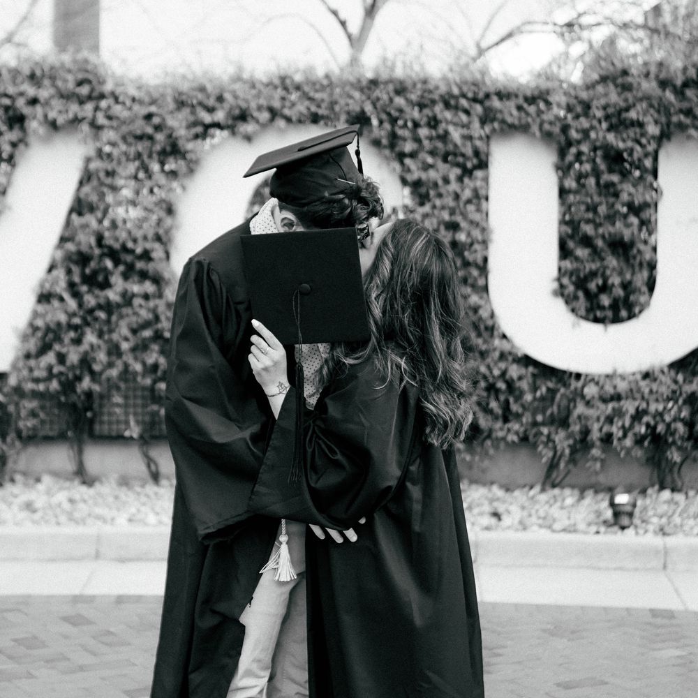 A graduate in cap and gown shares a romantic kiss with a partner outdoors near an ivy-covered wall.