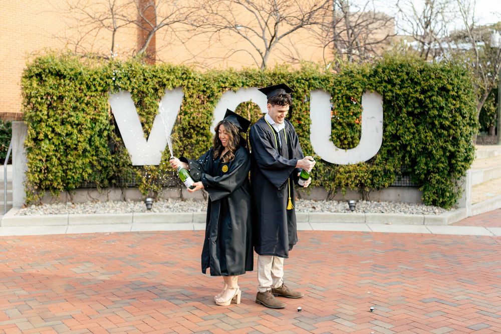 Two VCU graduates in black caps and gowns pose back-to-back in front of large VCU letter sign on brick plaza.
