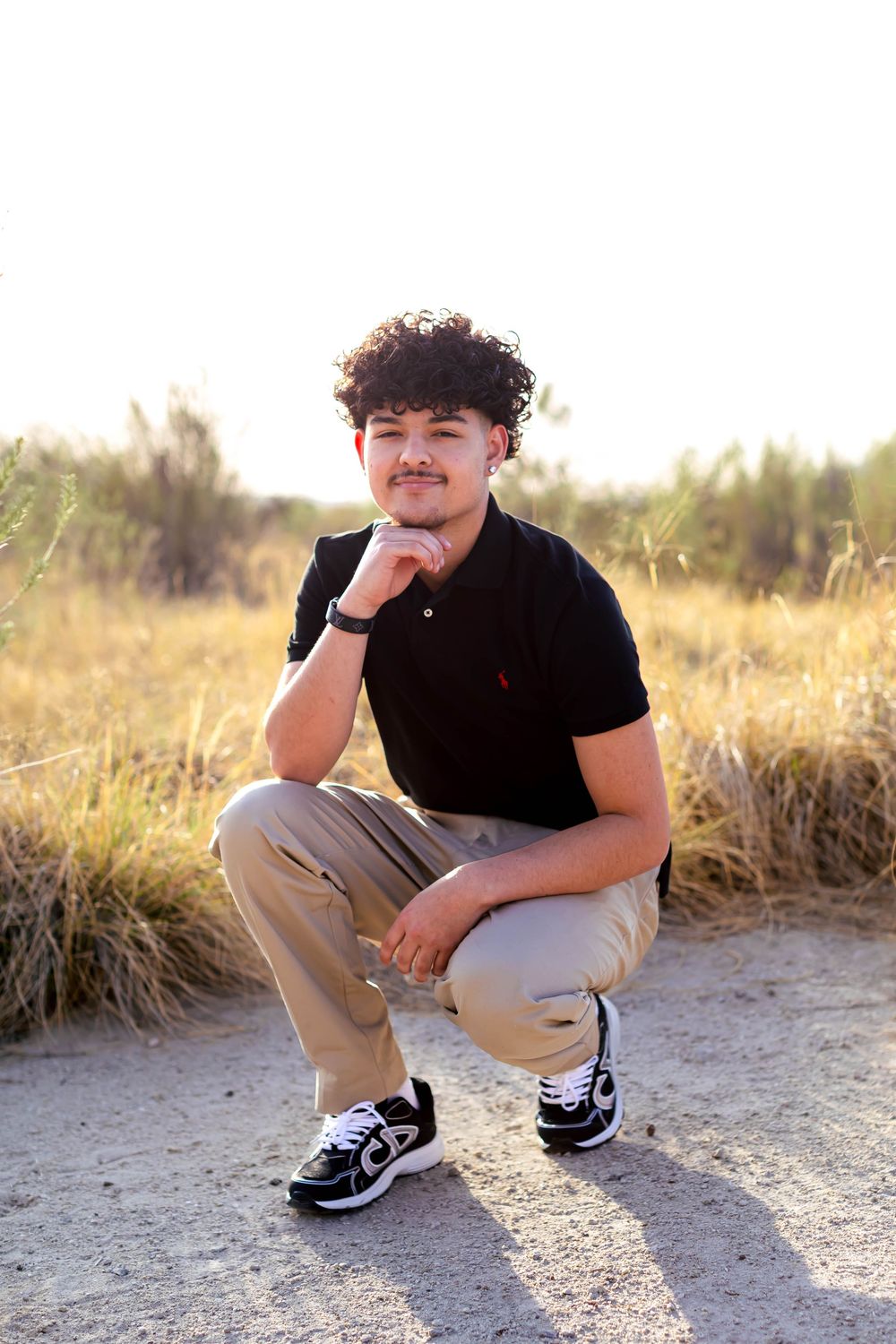 Young man with curly hair crouching outdoors in black polo and khaki pants, posing confidently in a sunny field.