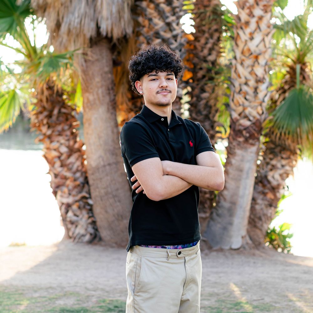 Young man with curly hair stands confidently with arms crossed near palm trees, wearing a black polo and khaki pants.
