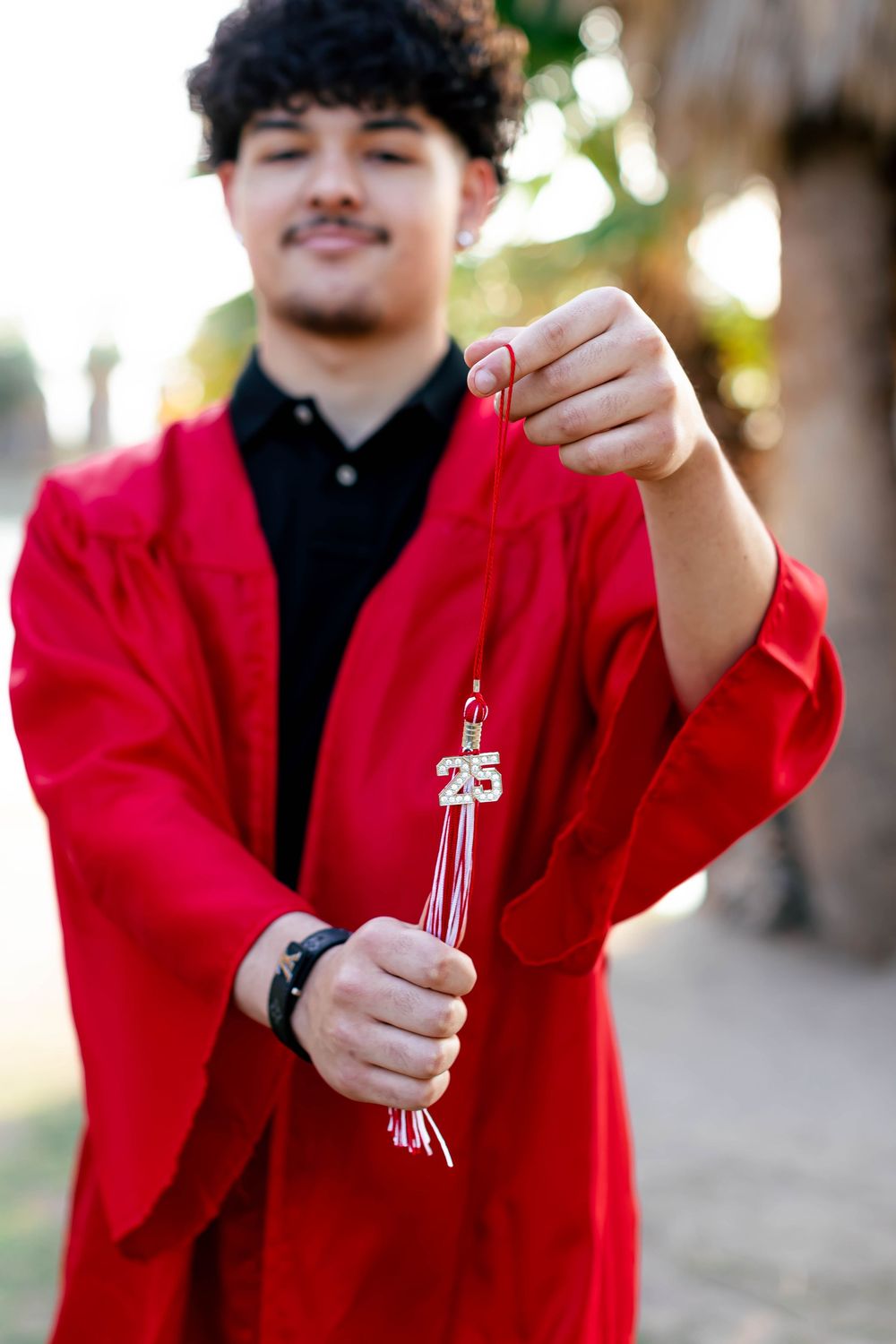 Young man in red graduation gown holding a Class of 2025 tassel outdoors, smiling proudly at his achievement.