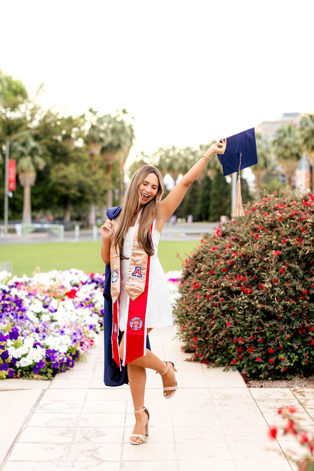 Smiling graduate in red sash holds cap up triumphantly beside colorful flowers at University of Arizona.