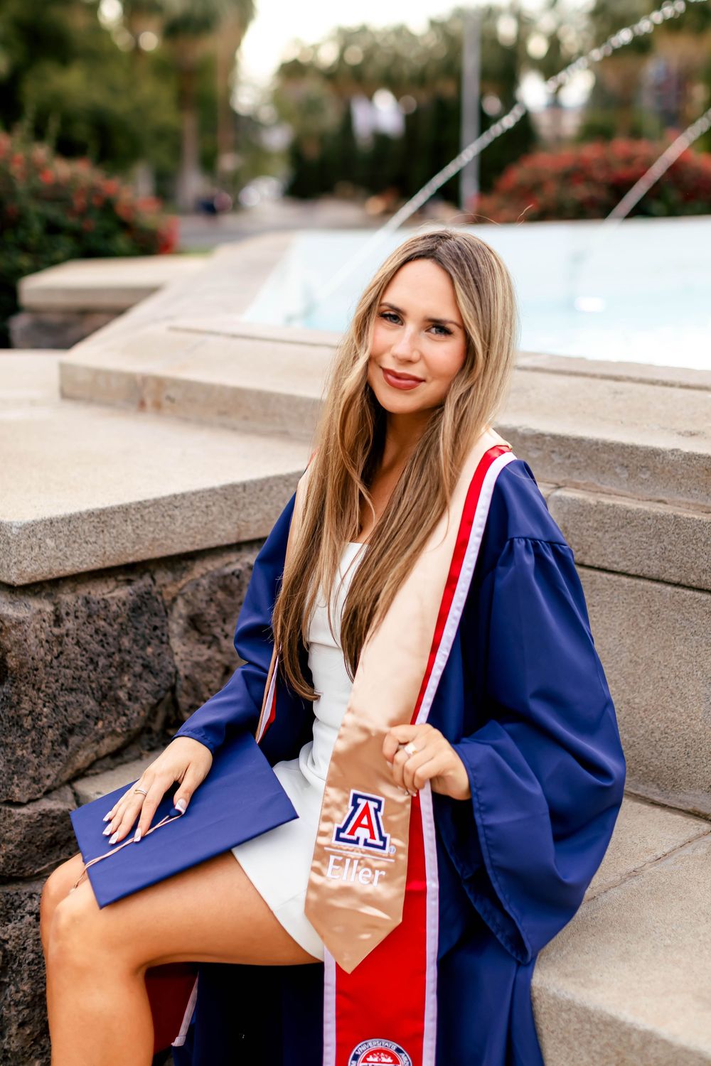 University of Arizona graduate in blue cap and gown holding diploma, seated outdoors near fountain.