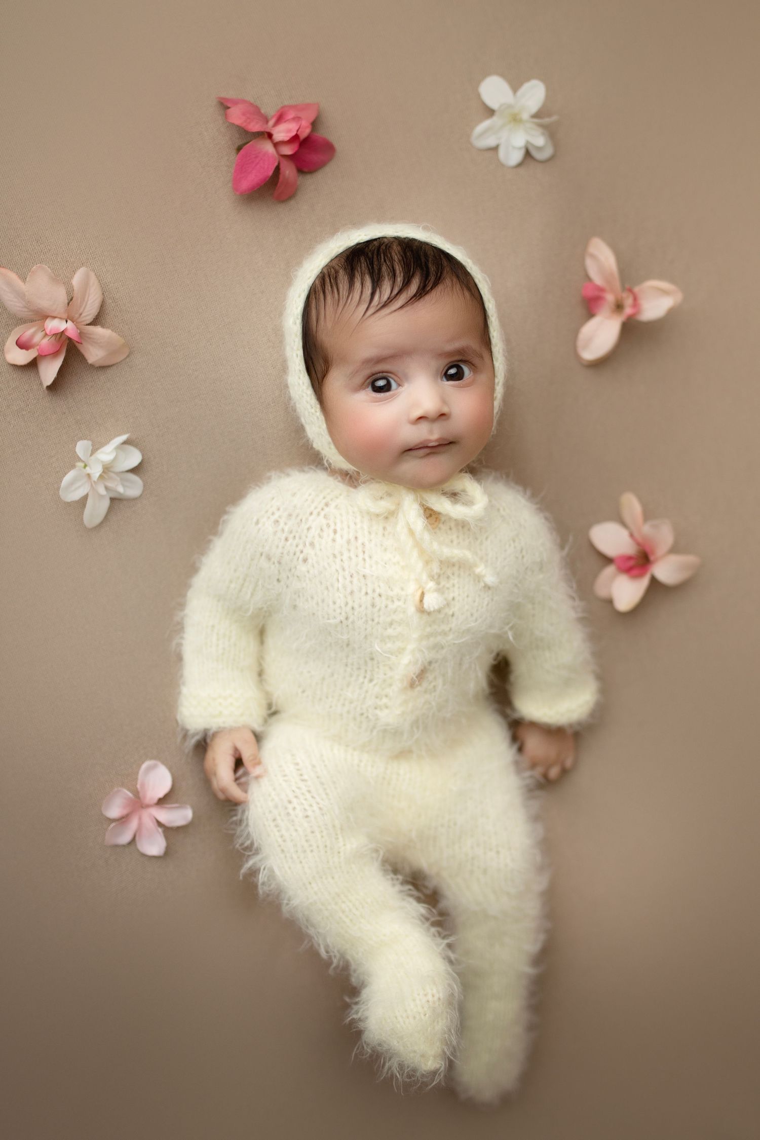 Baby in cream knit outfit surrounded by pink and white flower decorations.