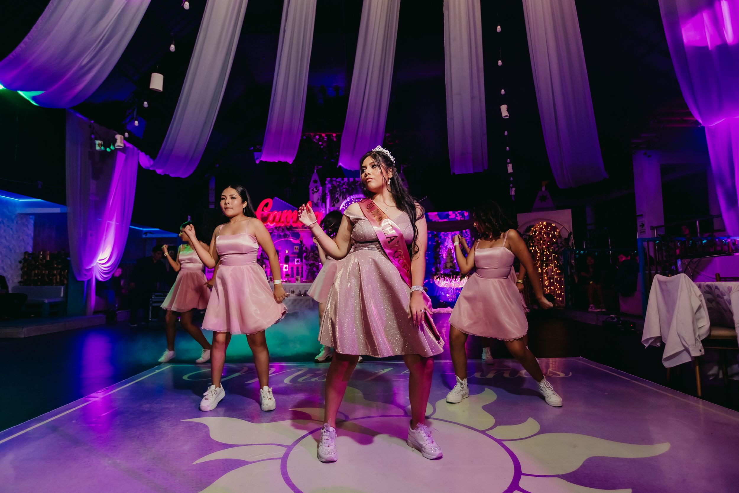 Dancers perform in pink dresses under purple lighting at a formal celebration.