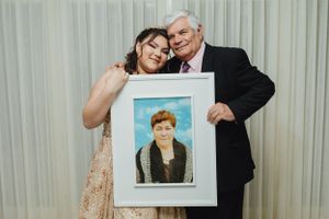 Two people at an event holding up a framed portrait photo against white curtained background.