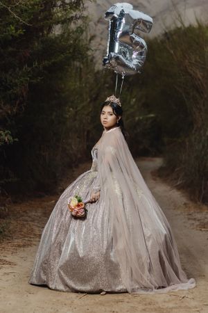 Person in silver quinceanera dress poses with number 15 balloon on outdoor path.