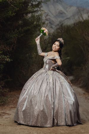 Person in shimmering silver ballgown poses dramatically with flowers on dirt path.