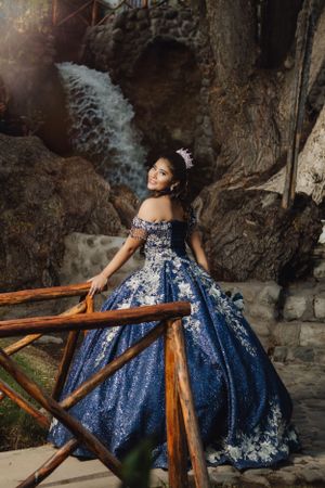 Person in ornate navy blue gown poses by waterfall with wooden railing.