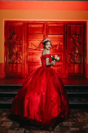 A formal portrait in a red ball gown against ornate red doors and wall decor.