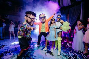 Energetic party scene with guests dancing in colorful outfits under neon lighting and smoke effects.