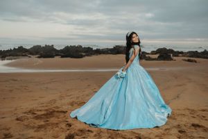 Person in light blue ball gown poses dramatically on beach at sunset.