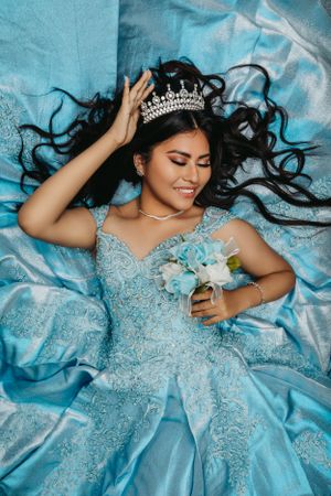 Overhead shot of person in turquoise quinceañera dress laying on matching fabric with tiara.