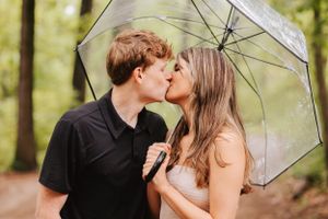 Romantic couple sharing a kiss under a clear umbrella in a wooded setting.