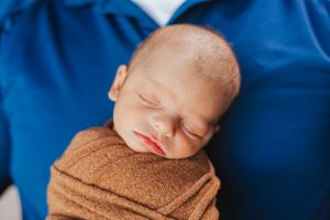 Newborn baby sleeping peacefully wrapped in brown swaddle against blue backdrop.
