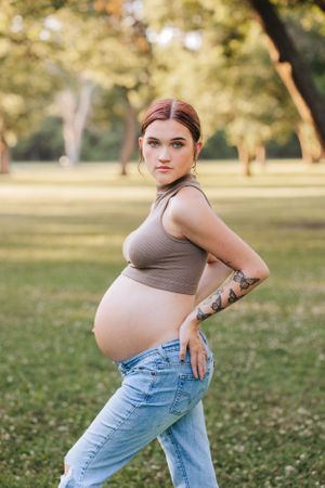 Expectant mother in casual attire poses outdoors in a sunny park setting.