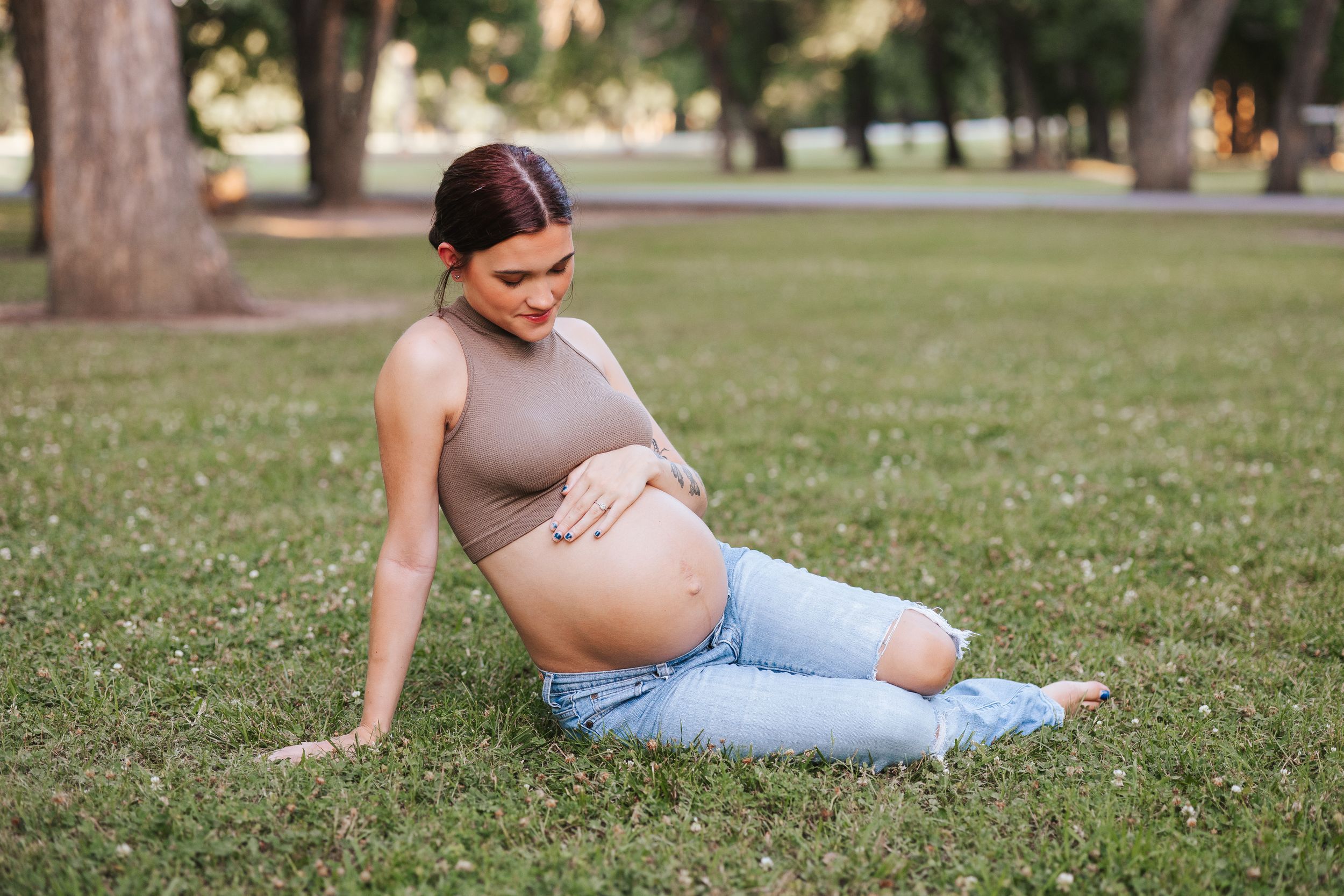 Pregnant woman in beige top and jeans relaxing on grass in park.