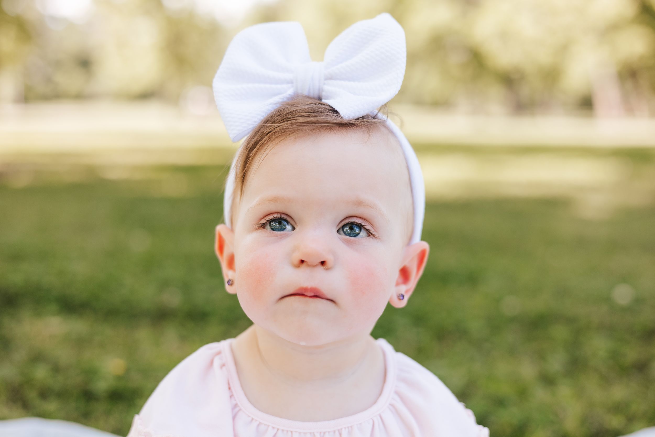 Baby wearing pink outfit and white bow headband on grass outdoors.