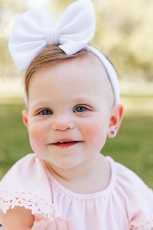 Close-up portrait of a baby wearing a white bow and pink dress smiling joyfully outdoors.