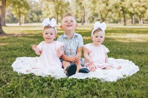 Children sitting together on white blanket in grassy outdoor setting.