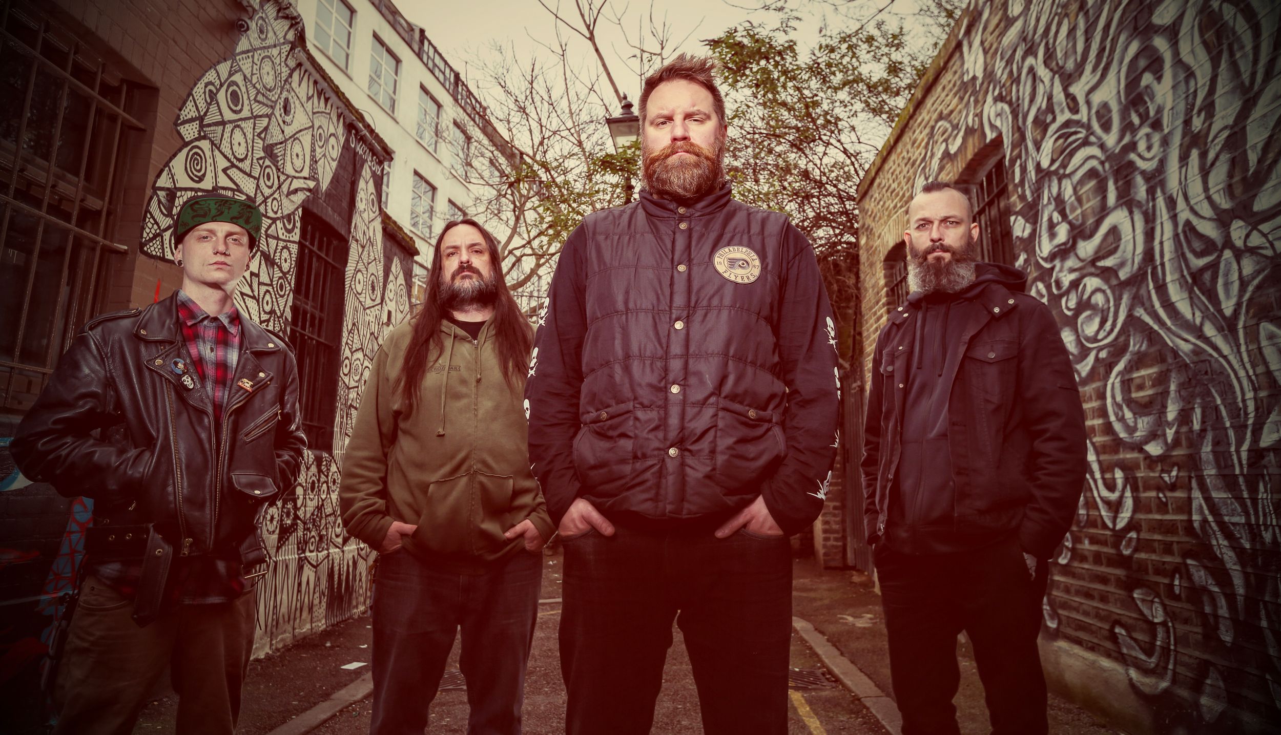 Four bearded men in dark jackets posing in front of graffiti wall for band photo.