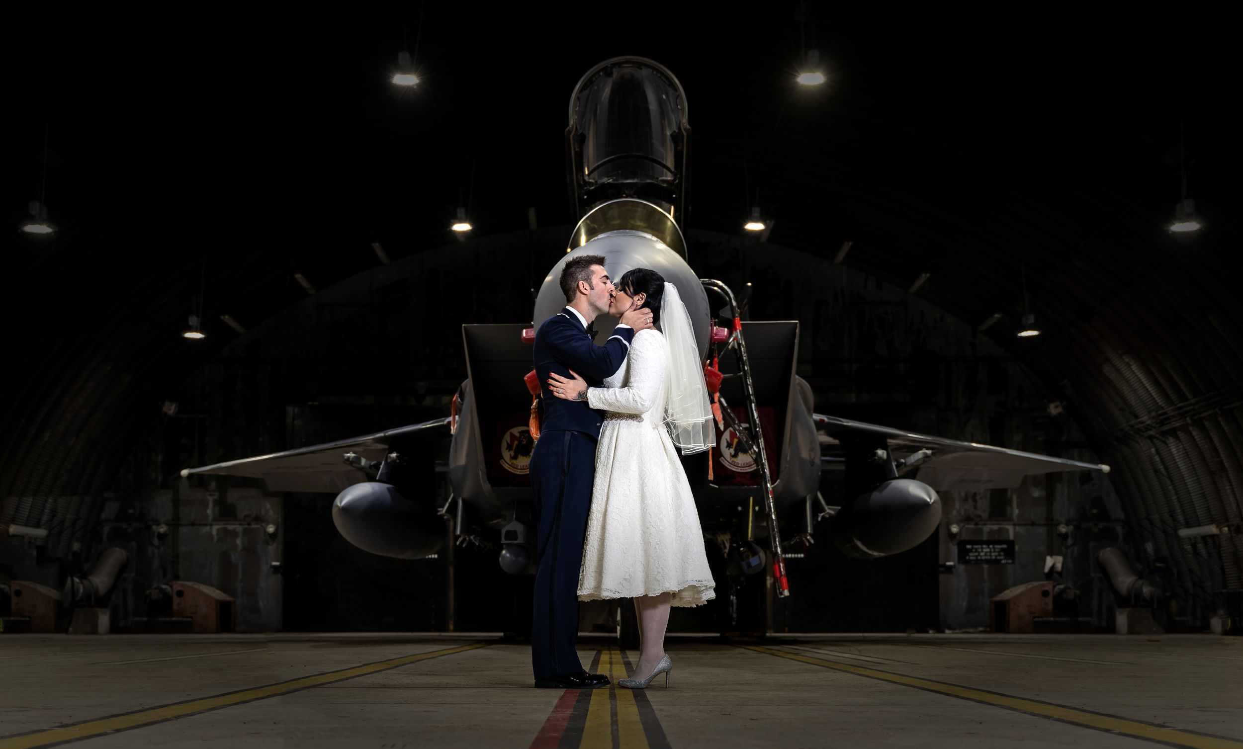 Couple sharing a kiss in front of a military jet aircraft inside a dark hangar.