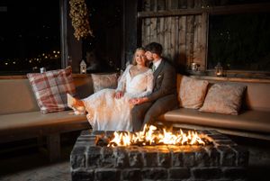 Bride and groom sitting together by an outdoor fire pit at a romantic night wedding.