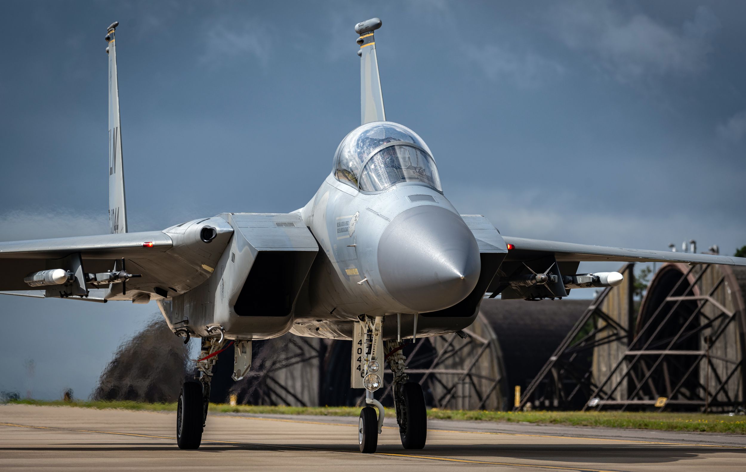F-15 fighter jet taxiing on runway with hangar in background on overcast day.