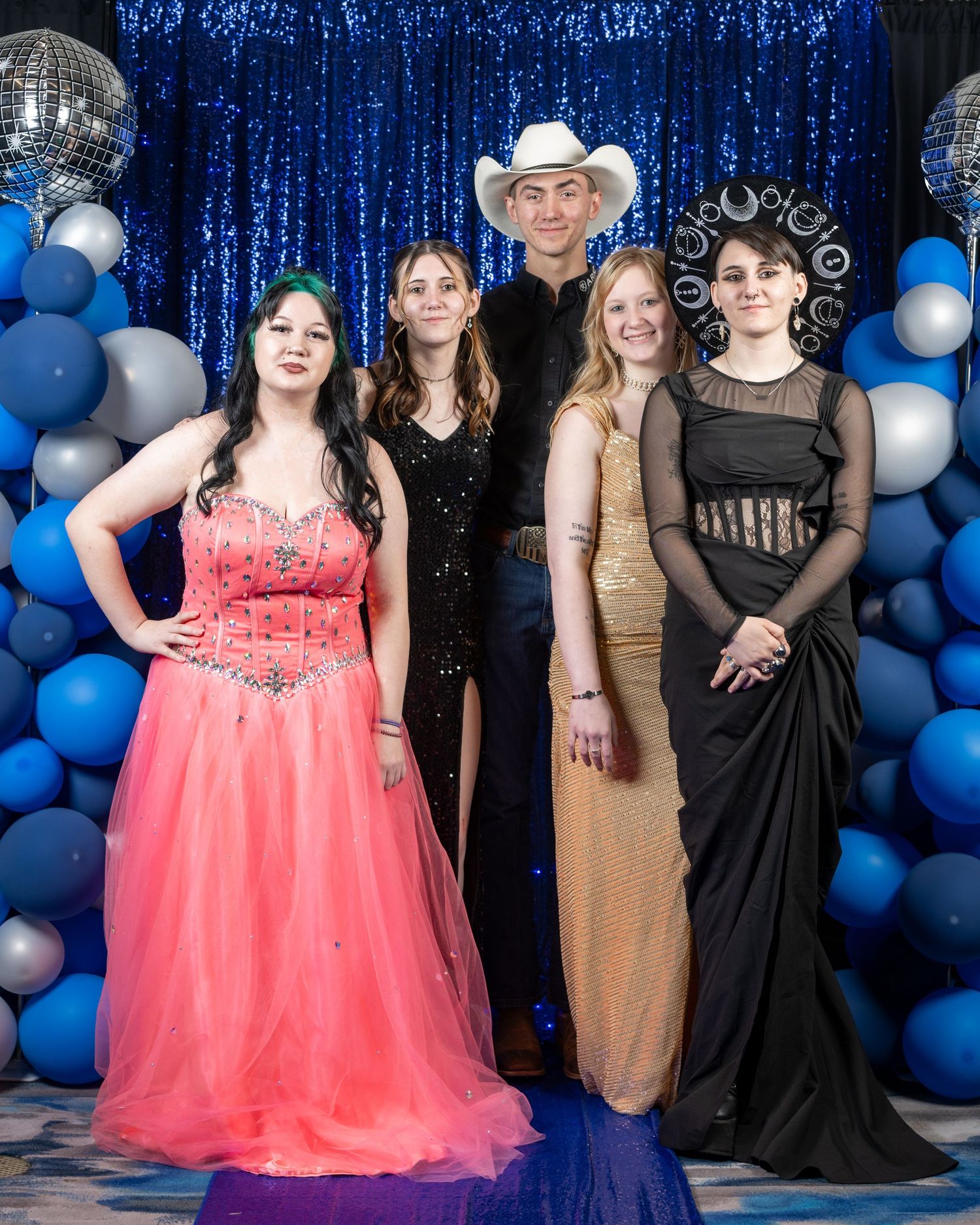 People at a formal event pose in elegant attire against a backdrop of blue and silver balloons and sparkly curtains.