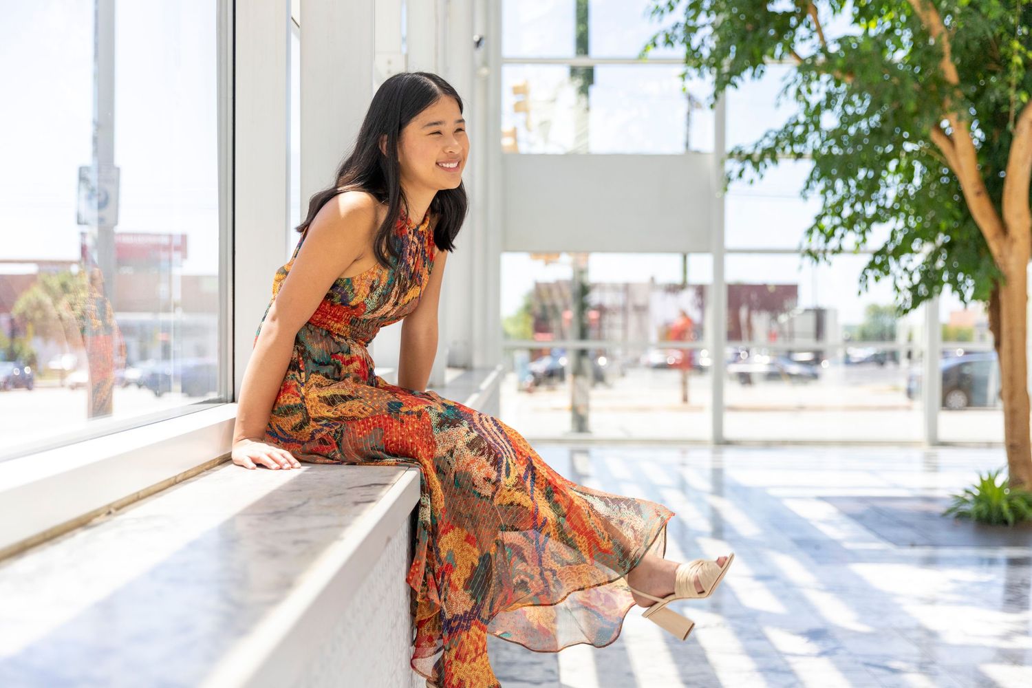 Someone in a colorful floral maxi dress sits casually on a white ledge in a bright outdoor space.