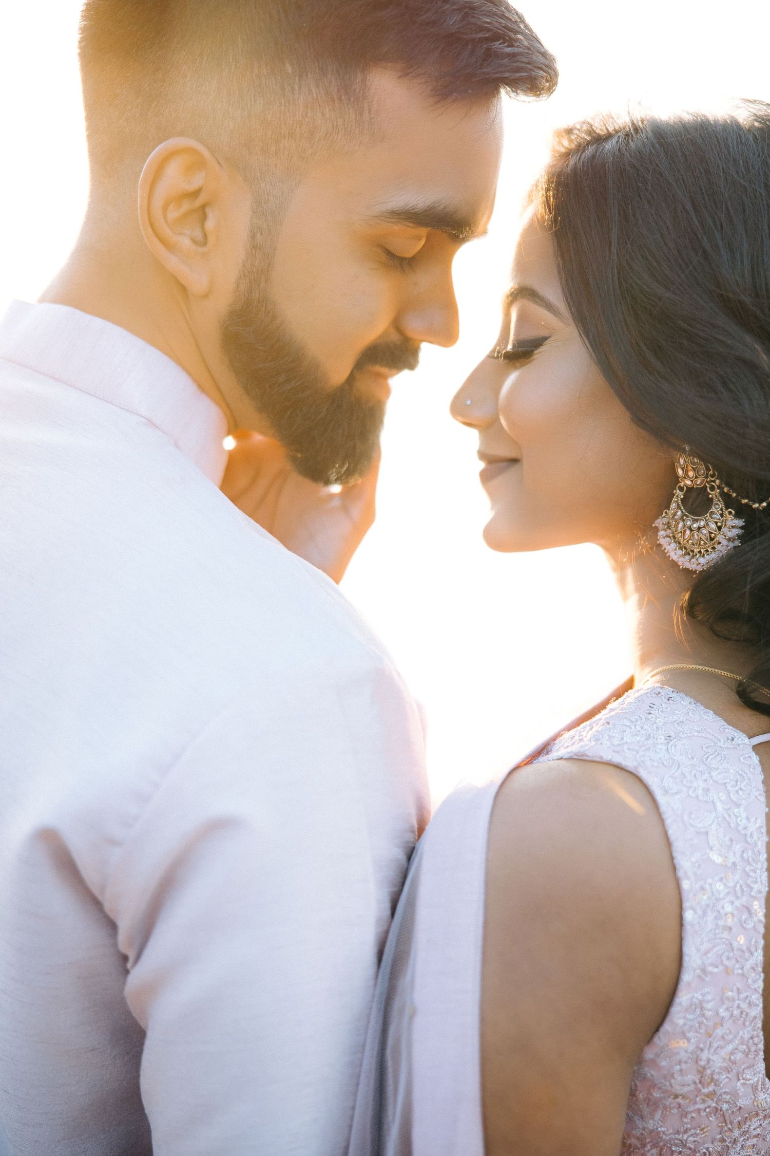 A romantic couple in white outfits sharing an intimate moment at sunset with golden backlighting.