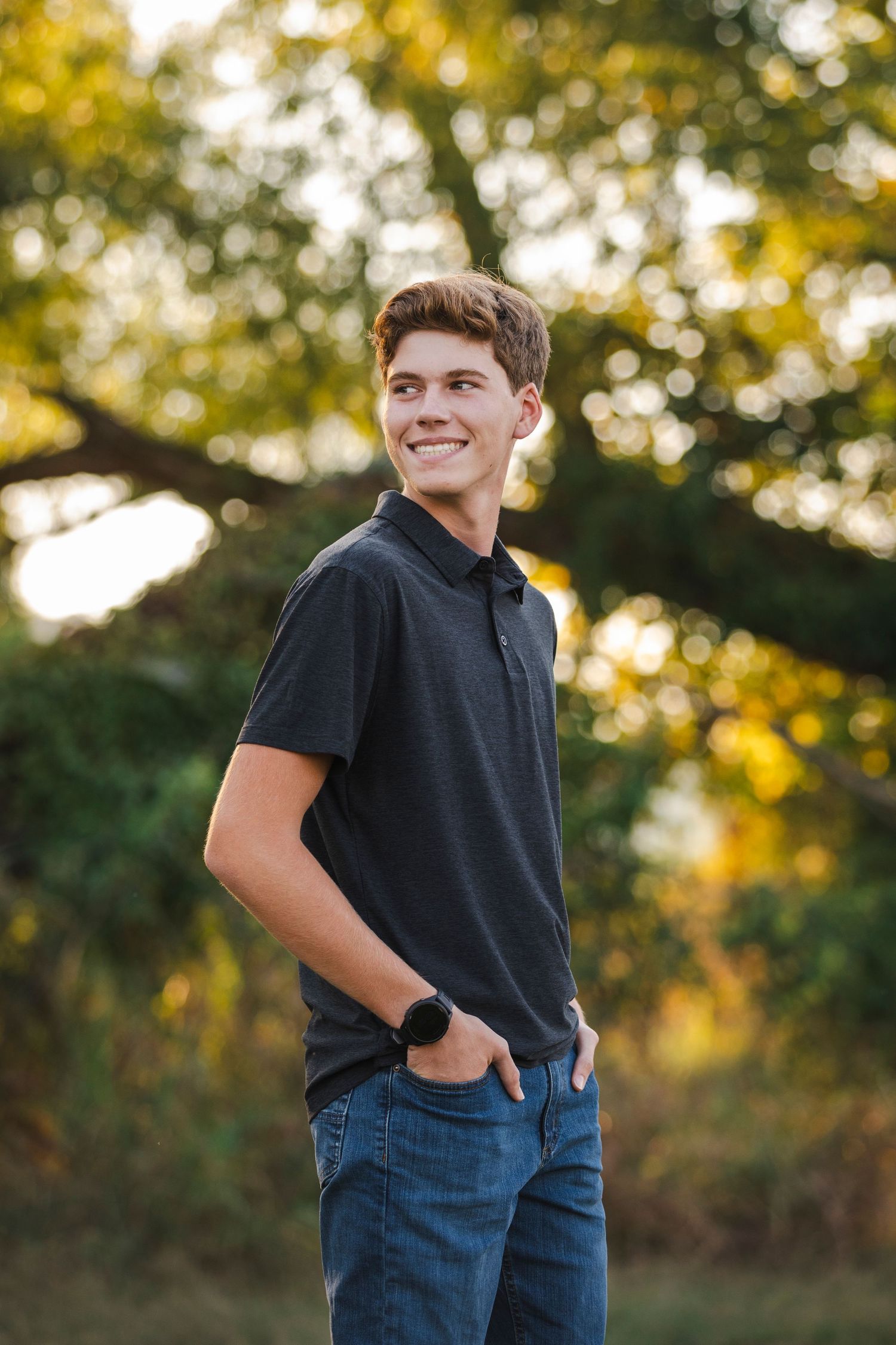 male student in black shirt in jeans with trees behind him