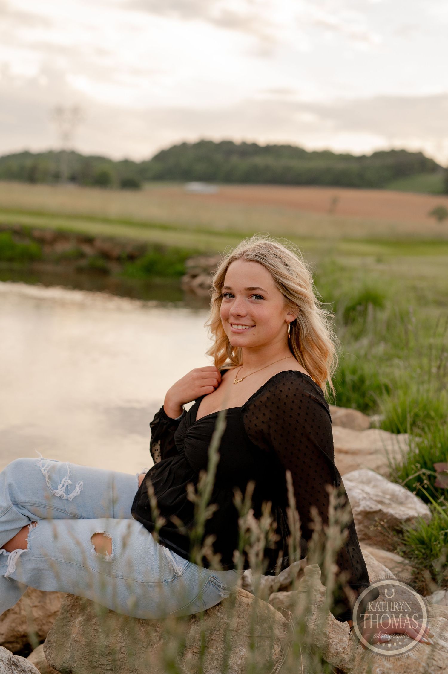 A sunset portrait in a field shows a casual pose against a scenic rural backdrop near a pond.