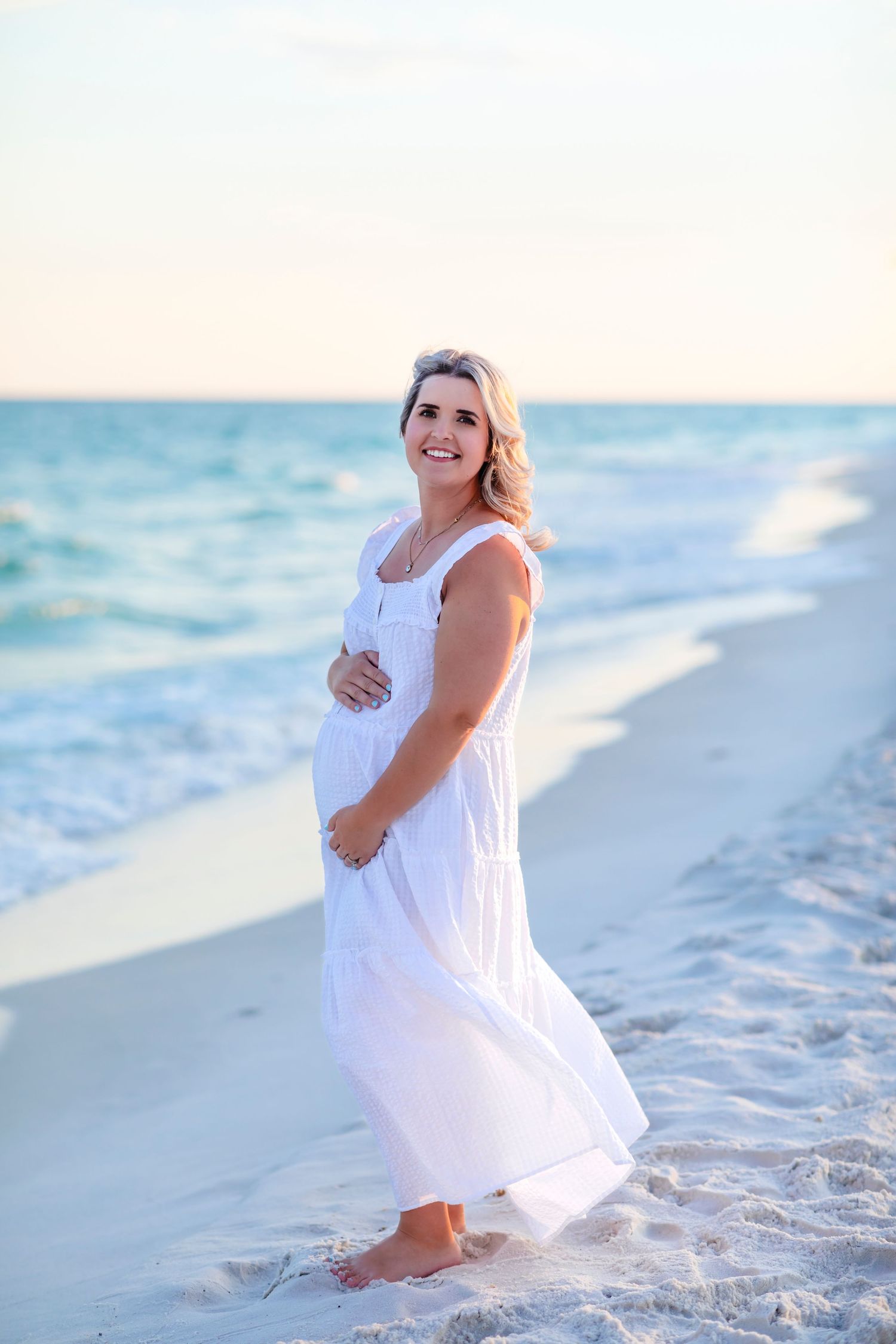 Expectant mother in flowing white dress poses on beach at sunset.