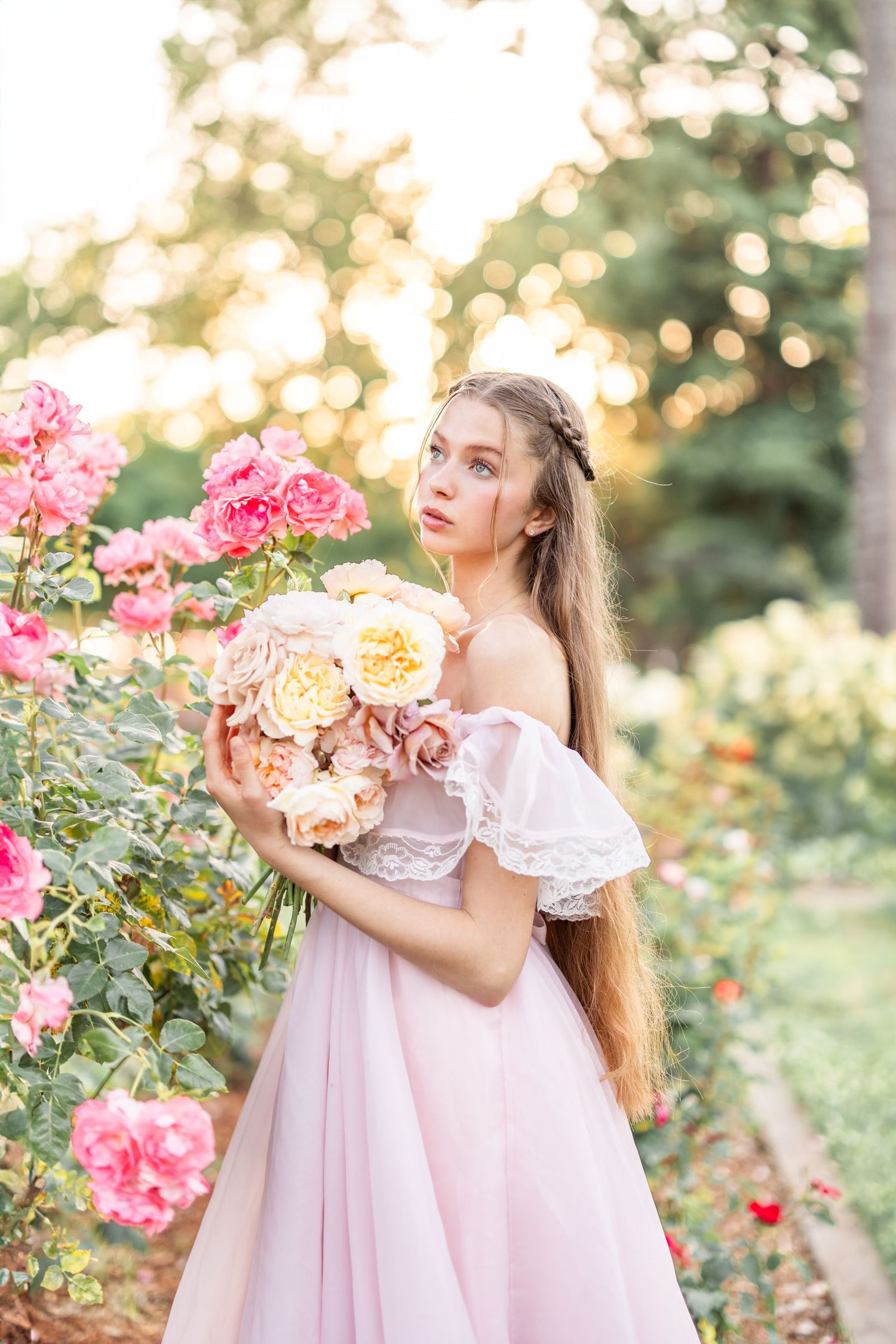 Young girl in pink dress poses with flowers in a blooming rose garden.