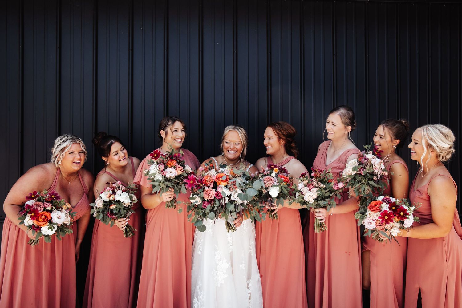A bridal party in coral dresses poses together against a dark backdrop with floral bouquets.