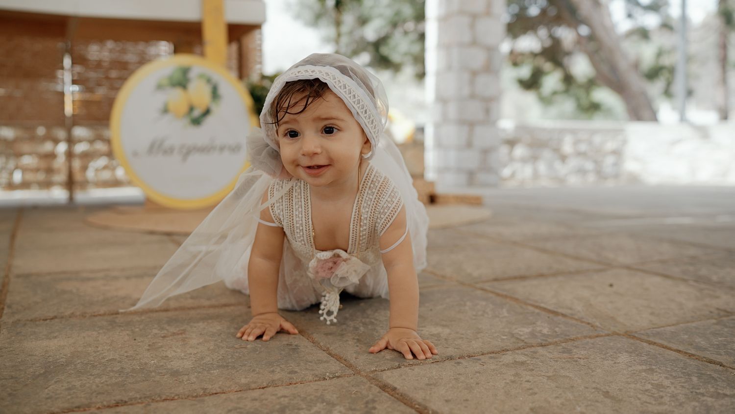 A baby girl in a white dress and veil crawls on stone tiles at an elegant outdoor baptism celebration.