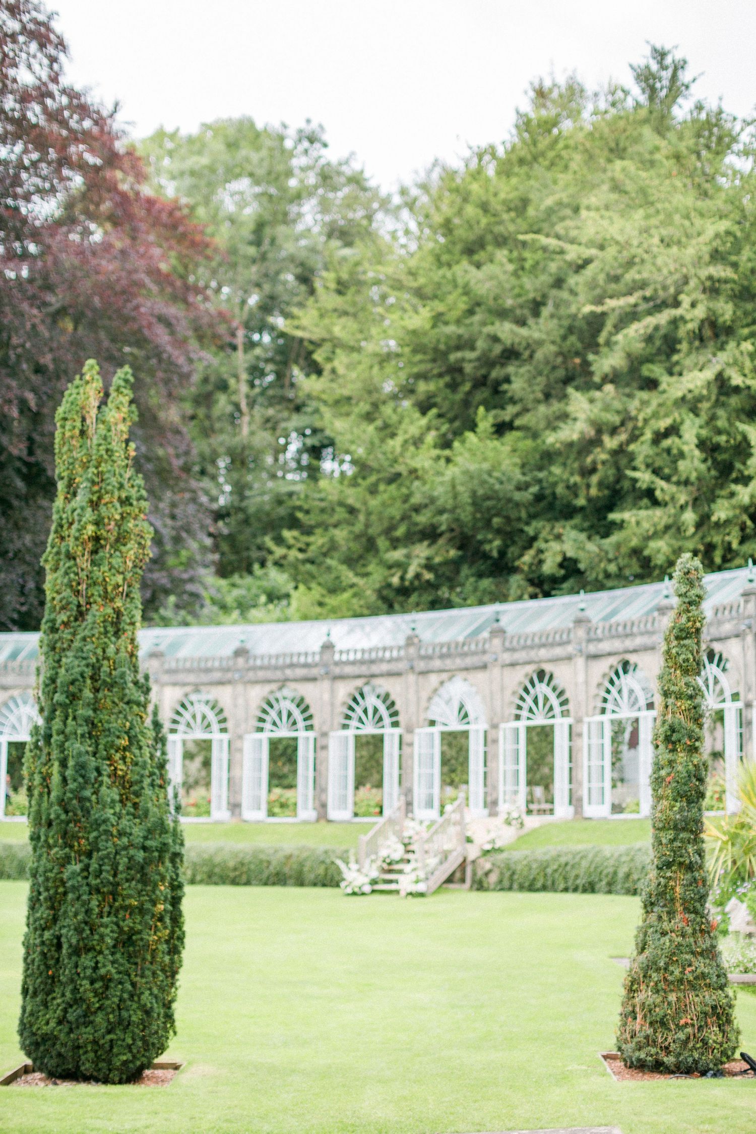 A series of cylindrical evergreen topiary trees line a manicured lawn with an ornate white balustrade wall in the background at sezincote