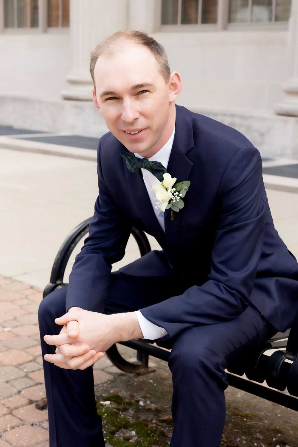 Groom in navy suit with boutonniere sitting on bench outdoors.
