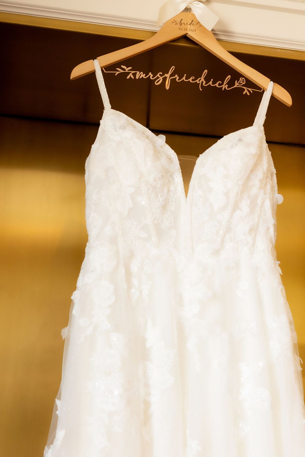 White lace wedding dress hanging on personalized wooden hanger against golden wall.