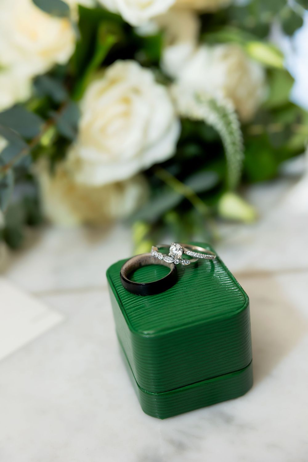 Diamond engagement ring and wedding band displayed on green velvet box with white roses in background.