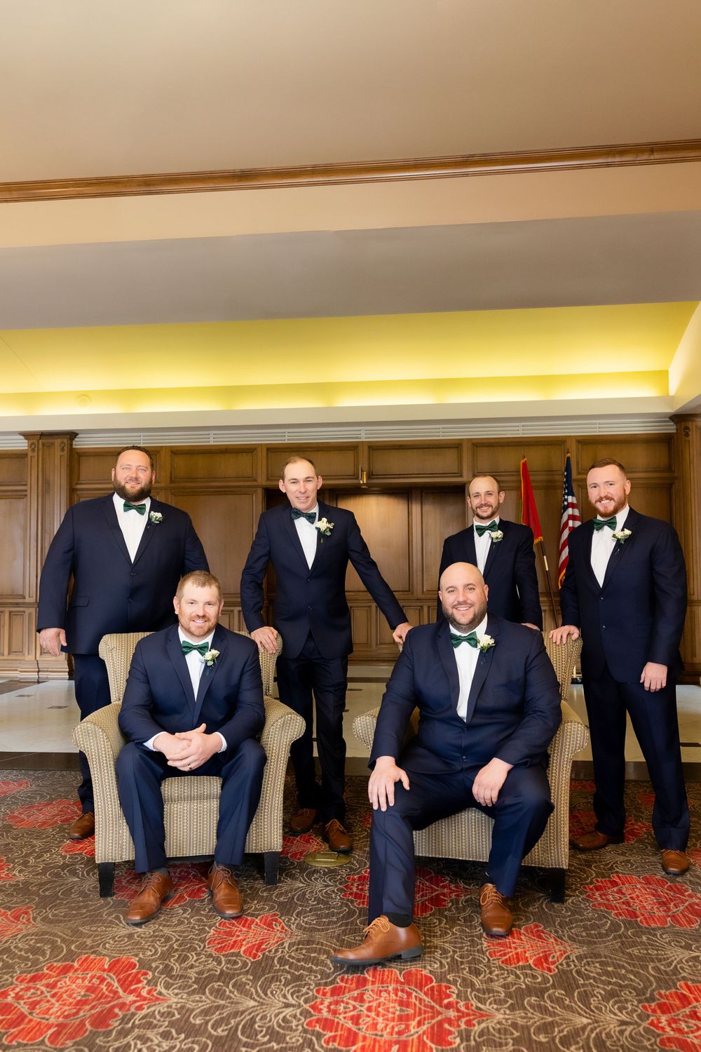 Wedding party groomsmen in navy suits and bow ties posed in formal hotel setting.