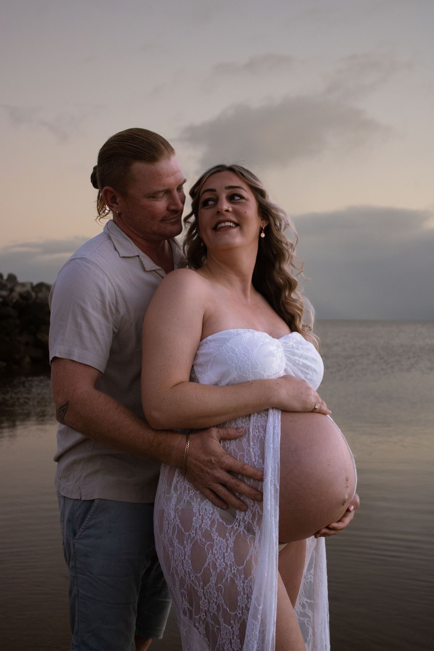 A couple shares an intimate moment during a maternity photoshoot at sunset by the water.