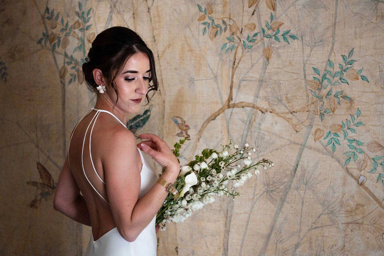 A bride in a white dress poses with white flowers against an ornate floral wallpaper backdrop.
