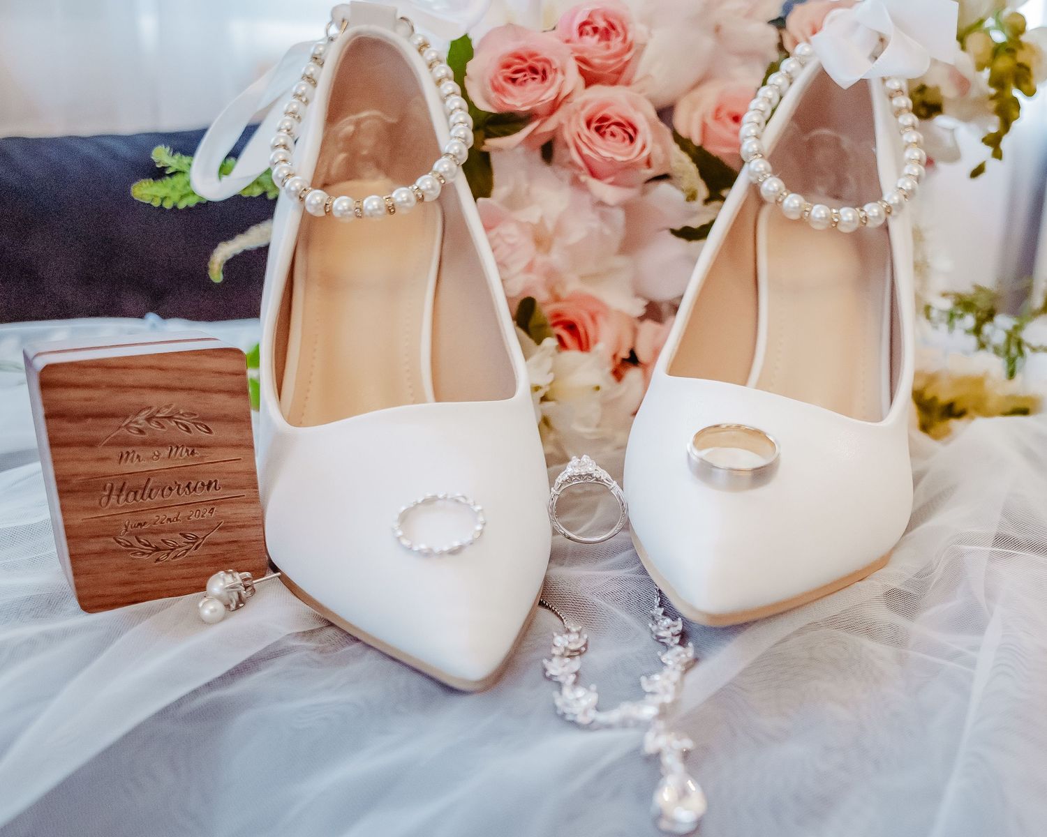 Elegant white bridal shoes with pearl straps displayed with pink roses and wooden box.