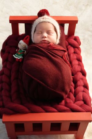 A sleeping baby swaddled in burgundy fabric resting on a small red wooden bench with a winter hat.
