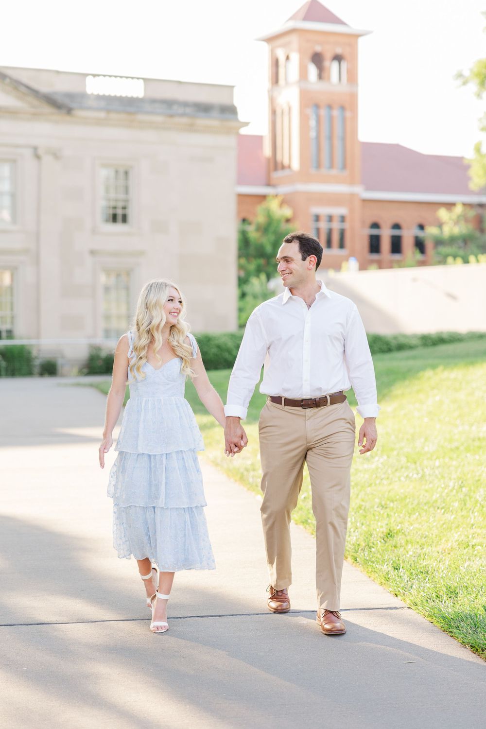 A couple takes a romantic stroll on a pathway with historic architecture in the background.
